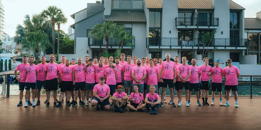 Team in pinken Shirts des BWT Best Water Run 2026 auf einer Holzterrasse vor einem Gebäude mit Palmen